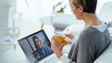 Person sitting on a couch holding a coffee cup while talking to a healthcare professional via a laptop