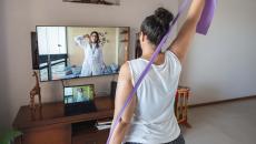 A woman watching and copying exercises with a resistance band in her living room, guided by a physical therapist online