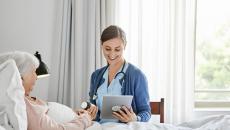 Person lying in bed with a healthcare provider sitting beside them in a chair holding a tablet