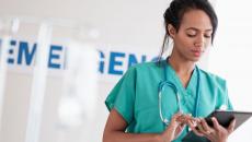 Healthcare provider wearing green scrubs and a stethoscope looking at a tablet in front of an emergency room sign