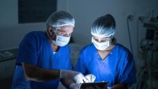 Two healthcare providers in scrubs wearing masks while in a dark room looking at a tablet
